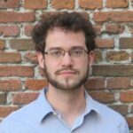 Headshot of a male with brown curly hair and scruffy beard wearing glasses.